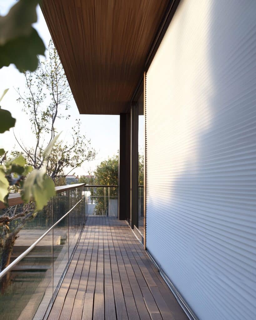 Balcon design bois et verre : espace extérieur moderne Terrasse en bois design, balustrade en verre, arbres luxuriants et store plissé blanc baigné de lumière naturelle.