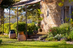 Maison en pierre avec pergola et guirlande lumineuse colorée. Jardin ensoleillé, grands pots en terre sur gazon fleuri de pâquerettes.