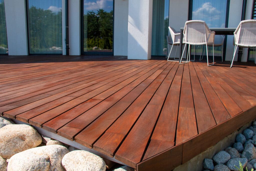 Terrasse en Bois Moderne : Aménagement Extérieur Élégant Grande terrasse moderne en bois avec mobilier de jardin devant une maison blanche et vitrée. Reflets d'arbres et ciel bleu.