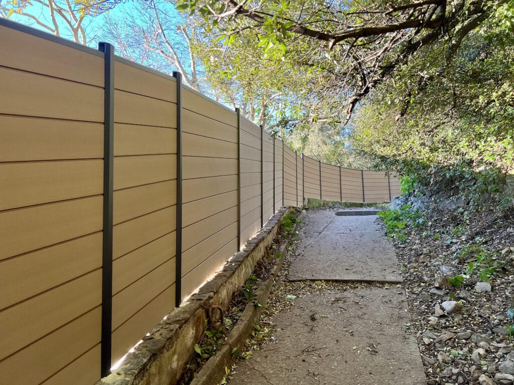 Clôture moderne en bois le long d'un sentier boisé ensoleillé Longue clôture moderne en bois clair avec poteaux sombres, serpentant le long d'un sentier extérieur bordé d'arbres et de verdure sous un ciel bleu.