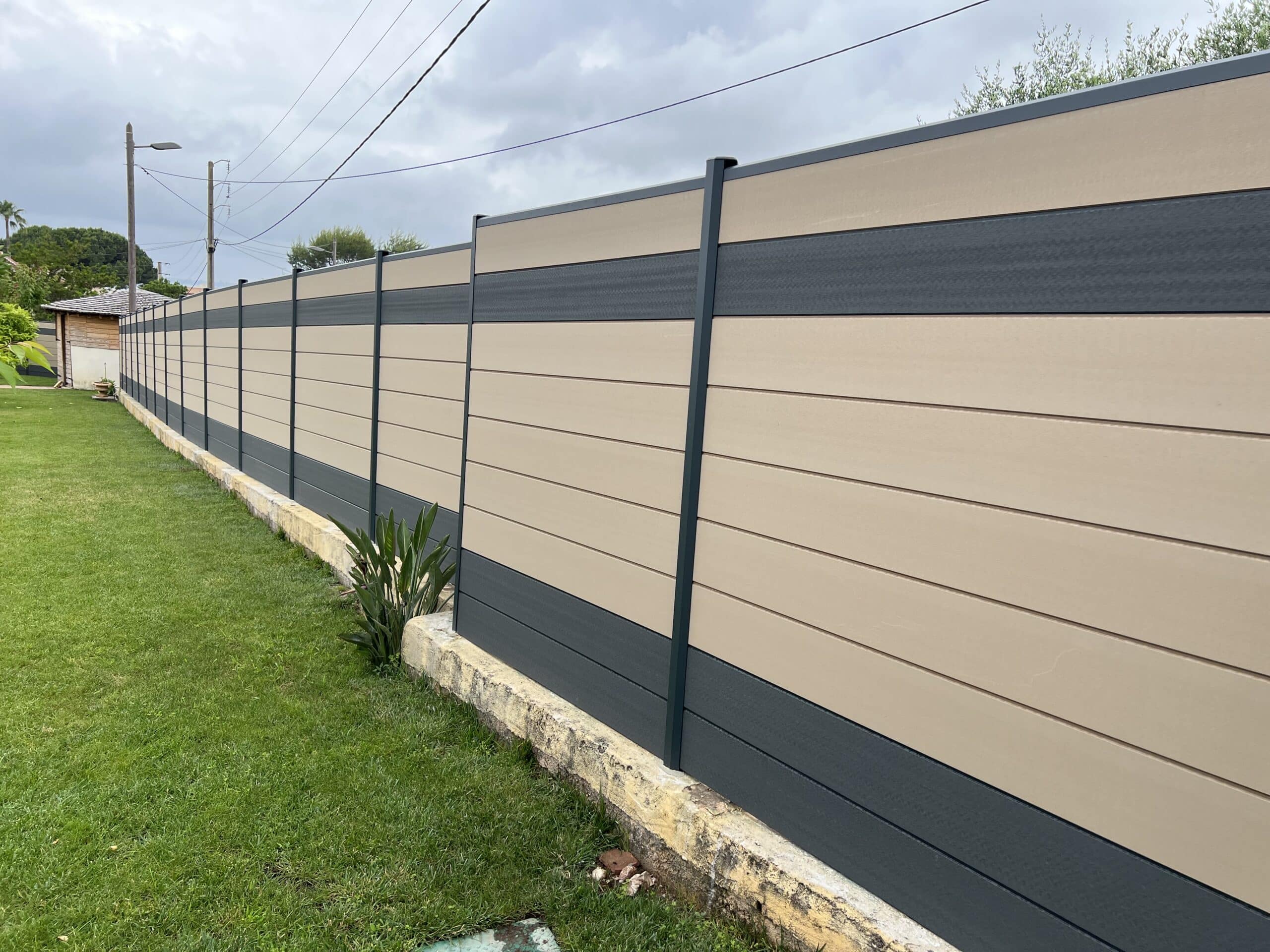 Longue clôture moderne en panneaux composites beige et gris anthracite, bordant un jardin vert avec un muret en béton sous un ciel nuageux.