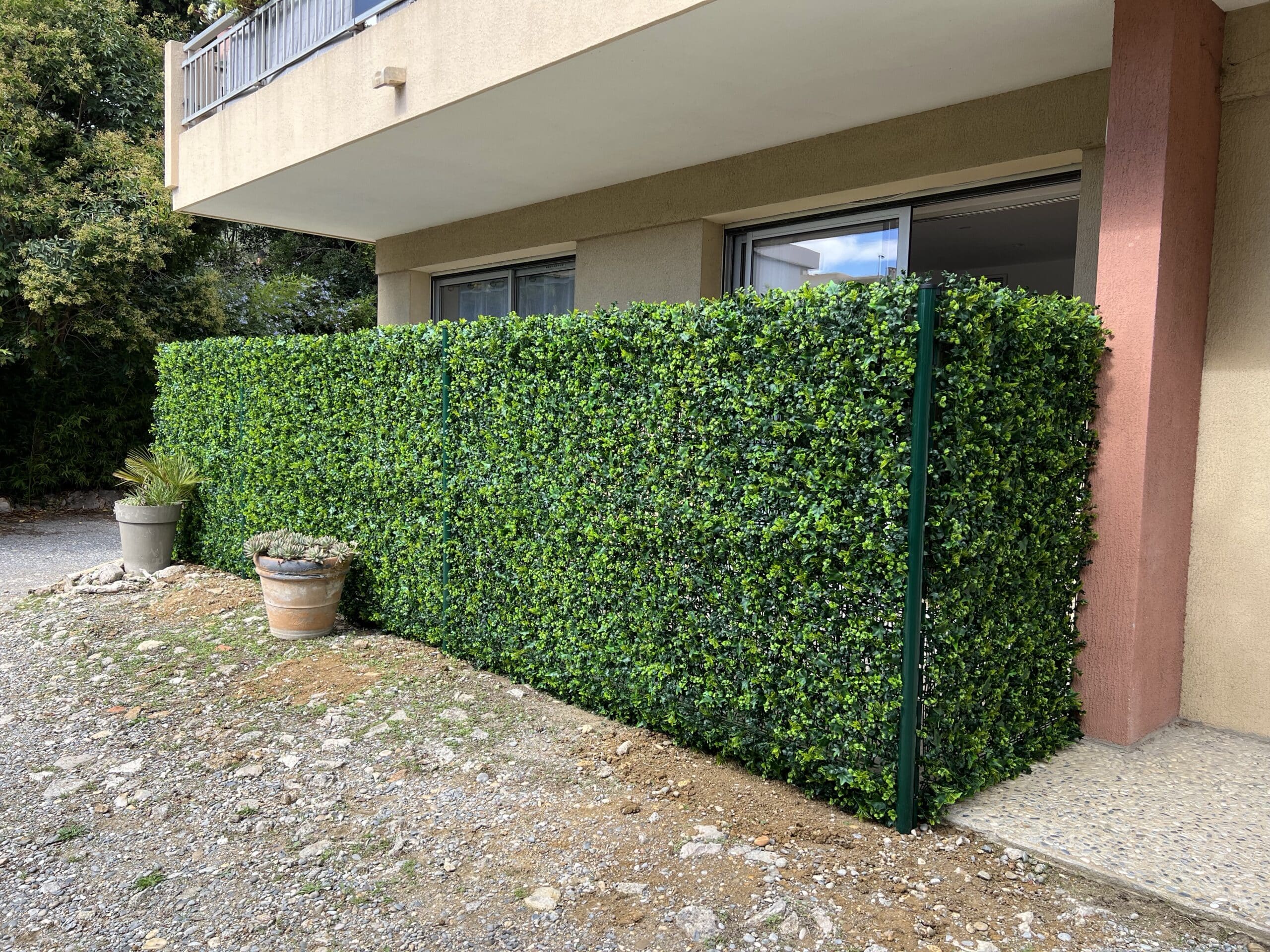 Longue haie artificielle verte brise-vue le long d'une terrasse d'appartement. Pots de fleurs, chemin de gravier et arbres luxuriants.