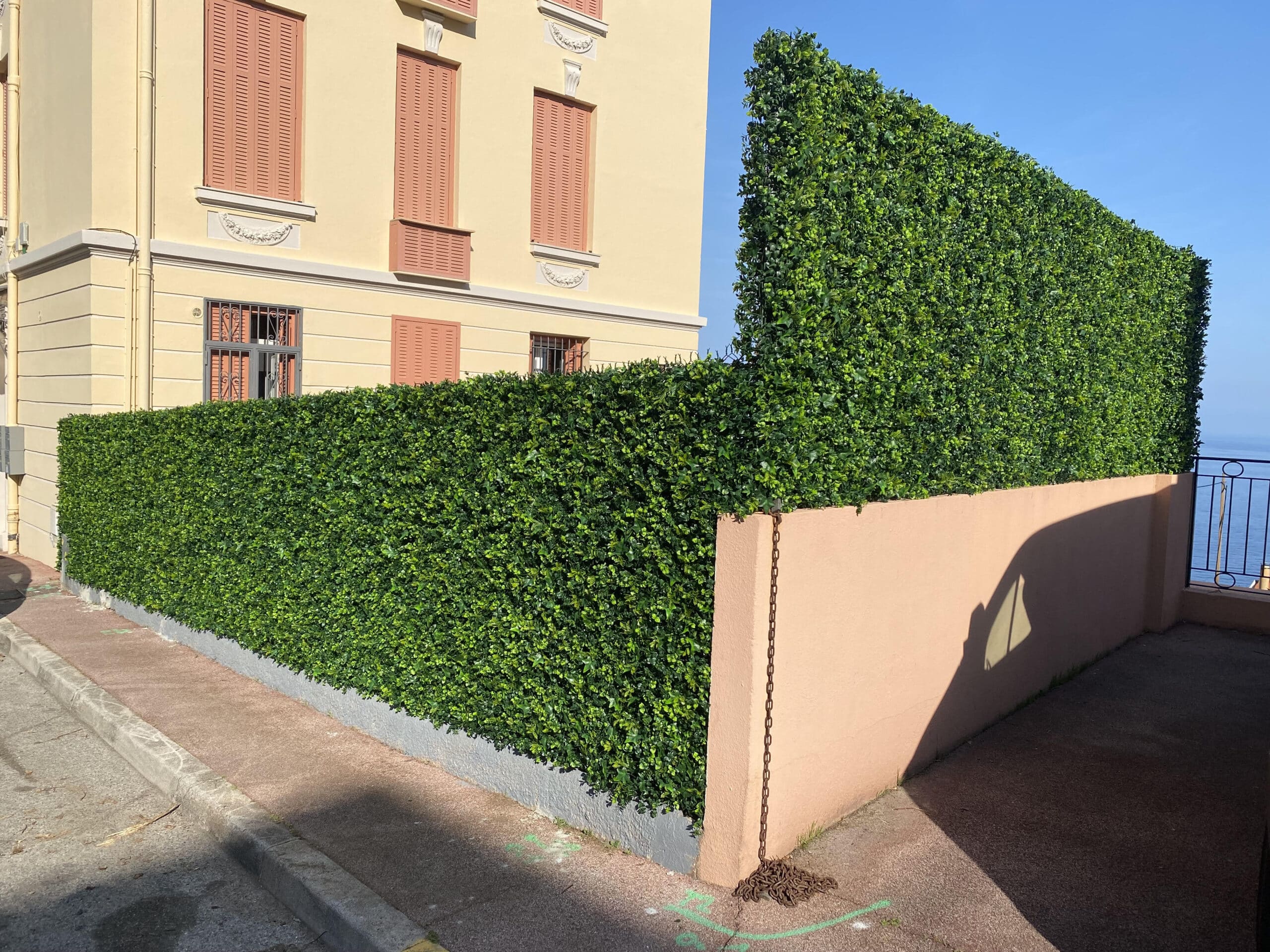 Façade jaune, volets corail, haie végétale artificielle dense. Mur rose avec chaîne rouillée sur trottoir. Mer Méditerranée et ciel bleu.