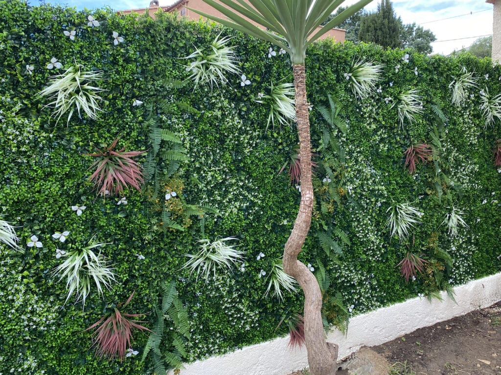 Mur végétal artificiel : Décoration de jardin sans entretien Mur végétal artificiel vert luxuriant avec fougères, plantes spikées et fleurs blanches. Tronc de Yucca courbé en premier plan.