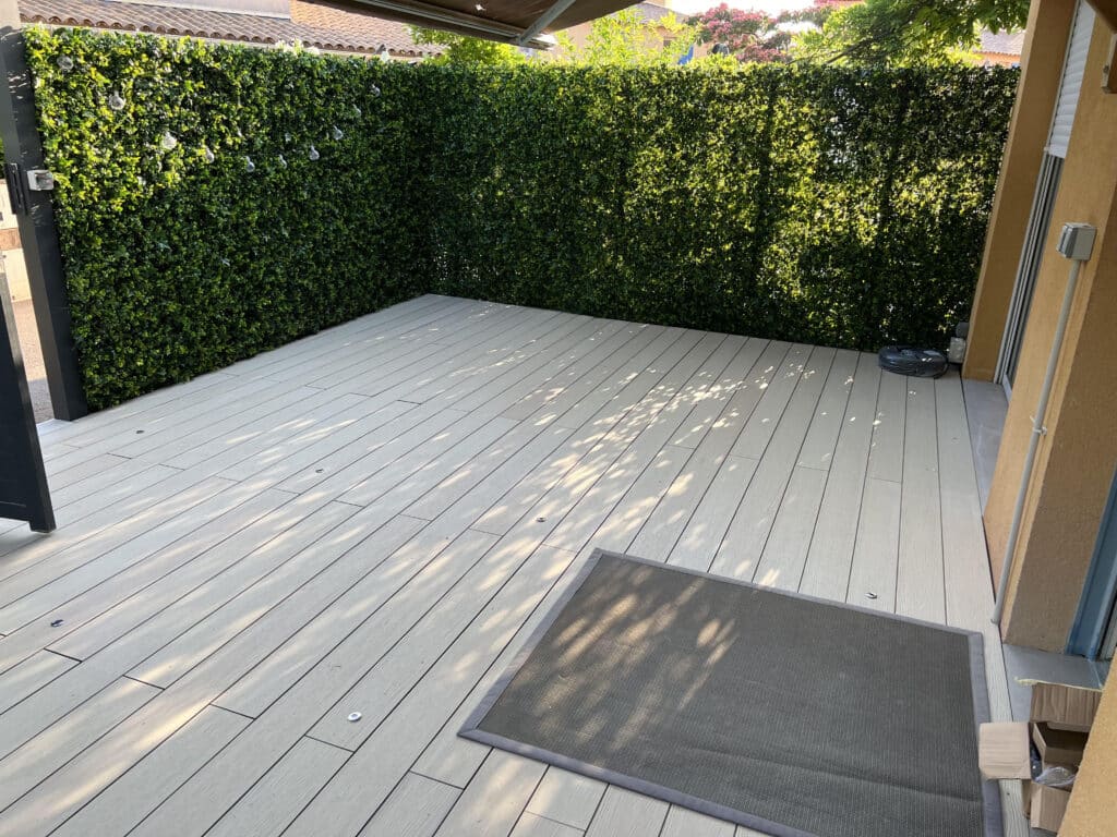 Terrasse moderne en composite: haie végétale et lumières Terrasse moderne en composite gris clair, haie artificielle verte avec guirlande lumineuse. Ombre d'arbres sur le sol, tapis gris extérieur.