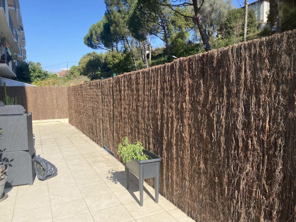 Aménager sa terrasse: clôture, salon de jardin et plantes Terrasse carrelée ensoleillée avec salon de jardin gris, grande clôture en brande, plantes vertes en pot et ciel bleu.